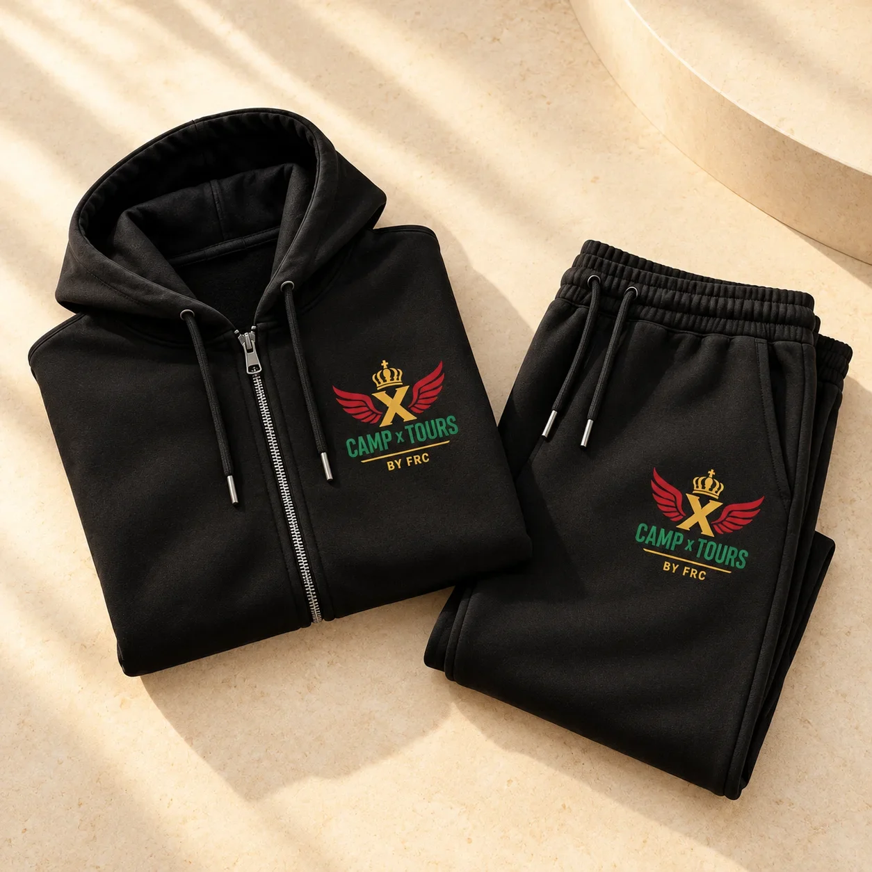 Black zip-up hoodie and joggers folded side by side on a cream surface, both showing the Camp X Tours by FRC emblem